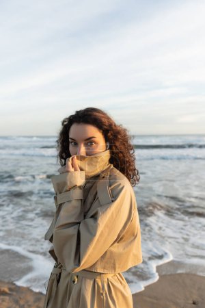 curly woman in wired earphones adjusting collar of beige trench coat and looking at camera near sea in Barcelona 