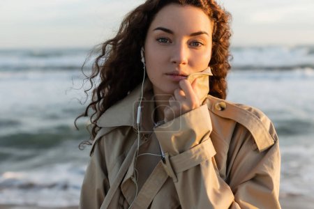 portrait of curly woman in beige trench coat and wired earphones listening music near sea in Barcelona 