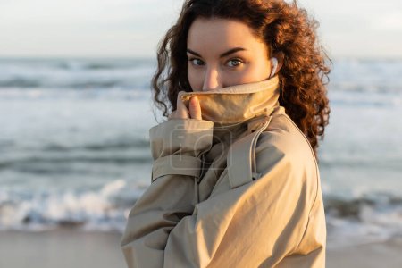 Téléchargez les photos : Portrait de femme bouclée dans des écouteurs filaires ajustant le col de trench-coat beige et regardant la caméra près de la mer - en image libre de droit
