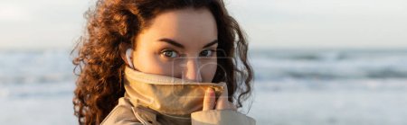portrait of curly woman in wired earphones adjusting collar of beige trench coat and looking at camera near sea, banner 