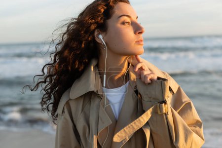 Curly woman in trench coat listening music in wired earphones on beach 