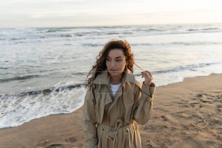 Junge Frau im Trenchcoat berührt lockiges Haar am Strand