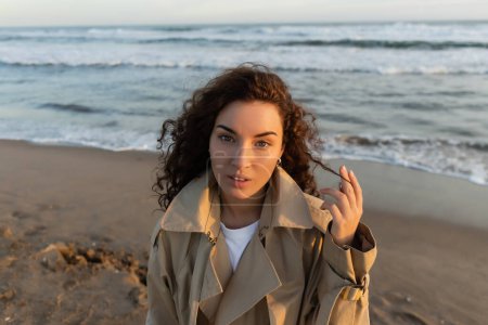 Foto de Curly woman in trench coat looking at camera on blurred beach near sea - Imagen libre de derechos