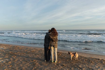 Back view of young women hugging near pug dog on beach in Spain 