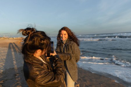 Young woman taking photo of cheerful friend on beach in Barcelona 