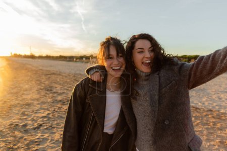 Cheerful curly woman hugging friend on beach during sunset 