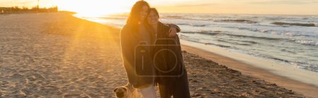 Smiling friends hugging near pug dog on beach in evening in Spain, banner 