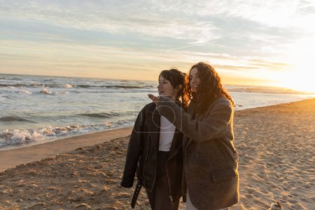 Positive Frau zeigt mit der Hand, während sie mit Freund am Strand am Meer spricht 