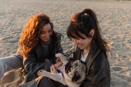 Fröhliche Frauen spielen mit Mops-Hund am Strand in Spanien 