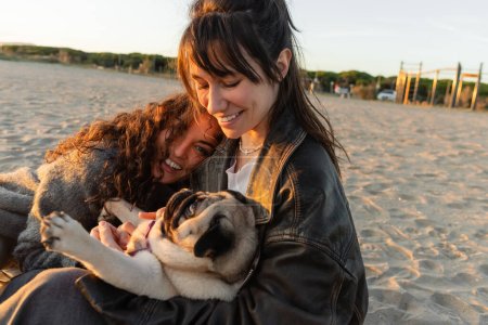Positive young women playing with pug dog on beach in evening 