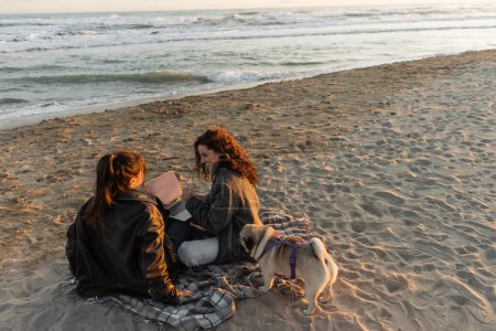 Lächeln Freunde mit Laptop in der Nähe Mops Hund am Strand in der Nähe des Meeres 