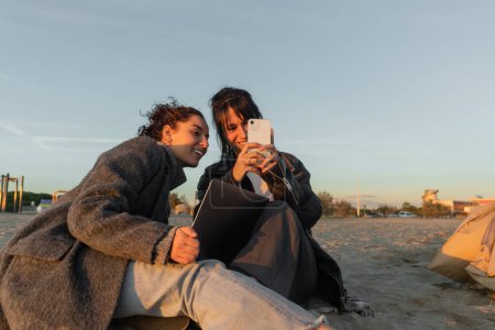 Smiling friends using smartphone and laptop on beach in Spain 