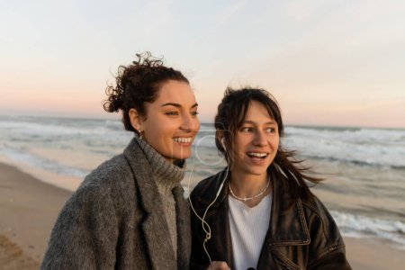 Téléchargez les photos : Des amis souriants écoutent de la musique dans des écouteurs filaires sur la plage en soirée en Espagne - en image libre de droit