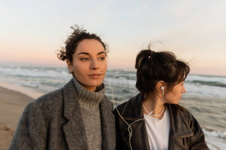 Téléchargez les photos : Femme bouclée écoutant de la musique dans des écouteurs filaires avec un ami sur la plage à Barcelone - en image libre de droit