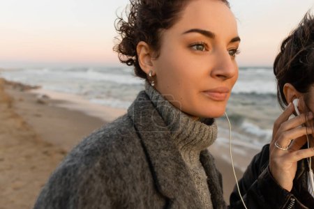Junge Frau hört mit Freund am Strand in Barcelona Musik in kabelgebundenen Kopfhörern 