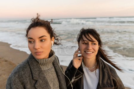 Smiling woman using wired earphones with friend on beach in Barcelona 