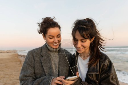 Positive friends in wired earphones using cellphone on beach in Spain 