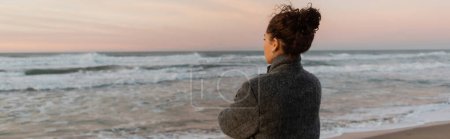 Curly woman in coat looking away while standing on beach near sea in Spain, banner 