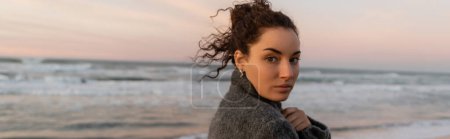 Téléchargez les photos : Jeune femme bouclée en manteau regardant la caméra avec la mer à l'arrière-plan, bannière - en image libre de droit