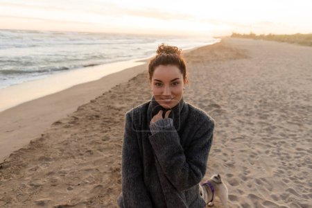 Positive Frau im Mantel blickt am Strand von Barcelona in die Kamera 