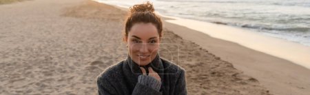 Smiling curly woman in coat looking at camera on beach in evening, banner 