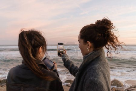 Curly woman taking photo on near friend with smartphone on beach in Barcelona 