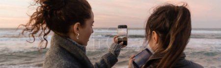 Friends taking photo on smartphones near sea during sunset, banner 