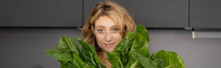 durchbohrte junge Frau lächelt und hält Grünkohlblätter in der Küche, Transparent 