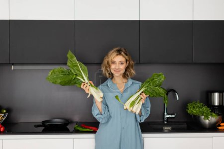 pierced young woman smiling and holding green cabbage leaves in kitchen 