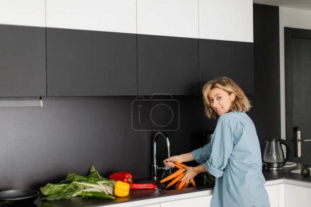 fröhliche junge Frau wäscht Möhren neben frischem Gemüse in der Küche 