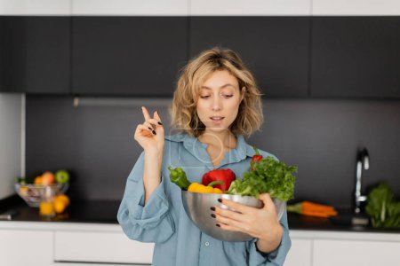 blonde young woman with wavy hair holding bowl with organic vegetables and showing idea sign 
