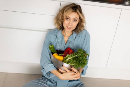 Photo for High angle view of cheerful young woman with wavy hair holding bowl with organic vegetables in kitchen - Royalty Free Image