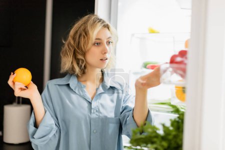 thoughtful young woman holding orange and looking at refrigerator in kitchen 