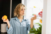 thoughtful young woman holding orange and looking at refrigerator in kitchen  Tank Top #637806078