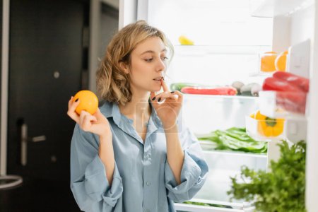 pensive young woman holding orange and looking at refrigerator in kitchen 