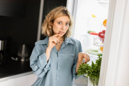 pensive young woman looking at camera near open refrigerator in kitchen 