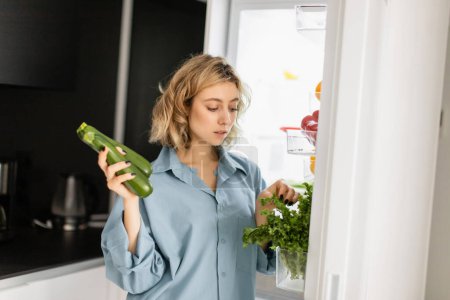 Foto de Mujer joven rubia en camisa azul mirando a la vegetación mientras sostiene calabacín cerca de nevera abierta en la cocina - Imagen libre de derechos