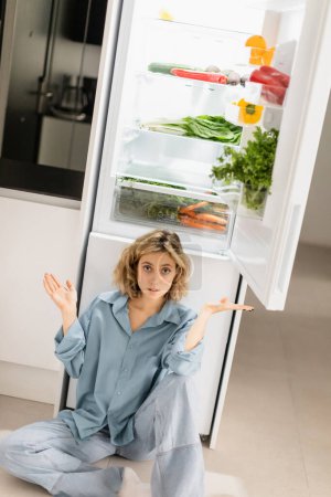 Foto de Mujer joven confundida sentada cerca de refrigerador abierto con productos frescos en la cocina - Imagen libre de derechos