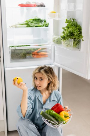 junge Frau sitzt neben geöffnetem Kühlschrank und hält Schüssel mit frischem Gemüse in der Küche 