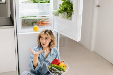 junge Frau sitzt neben geöffnetem Kühlschrank mit frischem Gemüse und wirft Zitrone in die Luft 