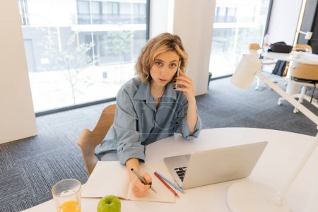 Blick aus der Vogelperspektive auf junge Frau, die in der Nähe von Laptop auf Schreibtisch mit Smartphone spricht 