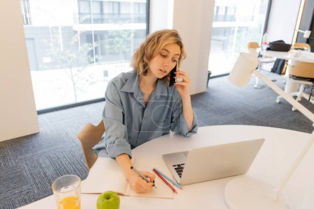 junge Frau mit welligem Haar spricht auf Smartphone neben Laptop auf Schreibtisch 