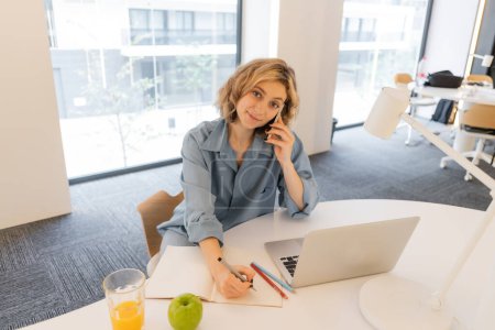 fröhliche junge Frau mit welligem Haar spricht auf dem Smartphone neben dem Laptop auf dem Schreibtisch 