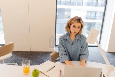Foto de Young woman with wavy hair using laptop near glass of orange juice and green apple on desk - Imagen libre de derechos