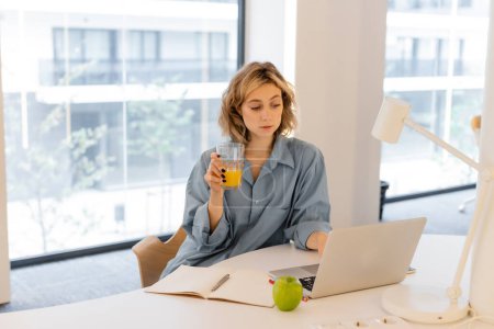 junge Frau mit welligem Haar hält Glas Orangensaft neben Laptop auf Schreibtisch 