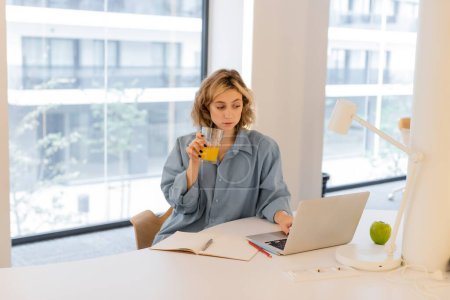 junge Frau mit welligem Haar hält ein Glas frischen Orangensaft in der Hand, während sie ihren Laptop benutzt 