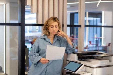 mujer rubia joven con cabello ondulado sosteniendo papel en blanco mientras está de pie cerca de la impresora en la oficina 