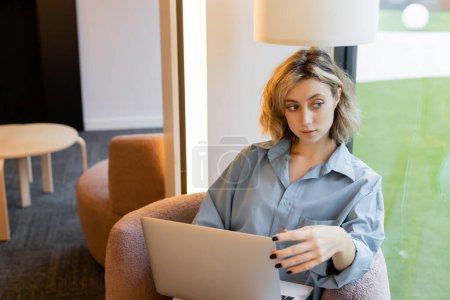 pensive and blonde woman with wavy hair looking away while sitting in armchair with laptop 