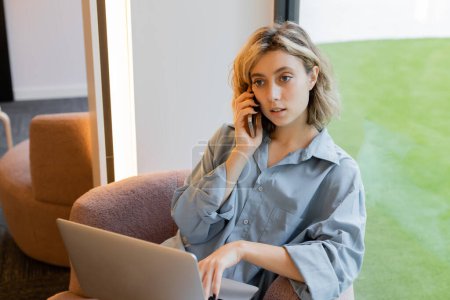 young blonde woman with wavy hair talking on smartphone while using laptop in cafe 