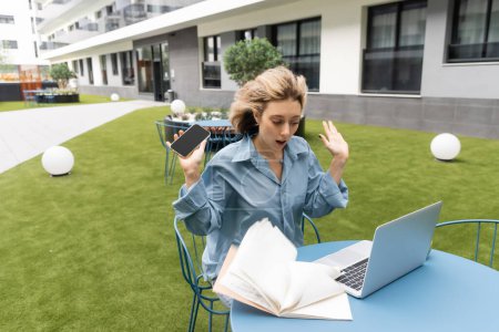 mujer conmocionada en camisa sosteniendo el teléfono inteligente mientras mira el viento soplando papeles de portátil cerca de la computadora portátil 